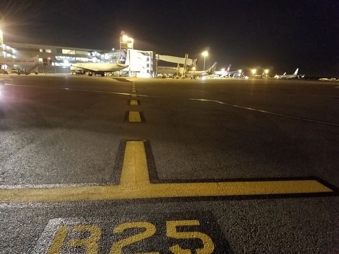 A runway with yellow lines and an airport in the background.